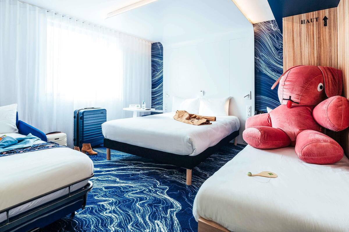 Habitación de hotel con dos camas, un peluche grande, una maleta y un muro de madera con un letrero.