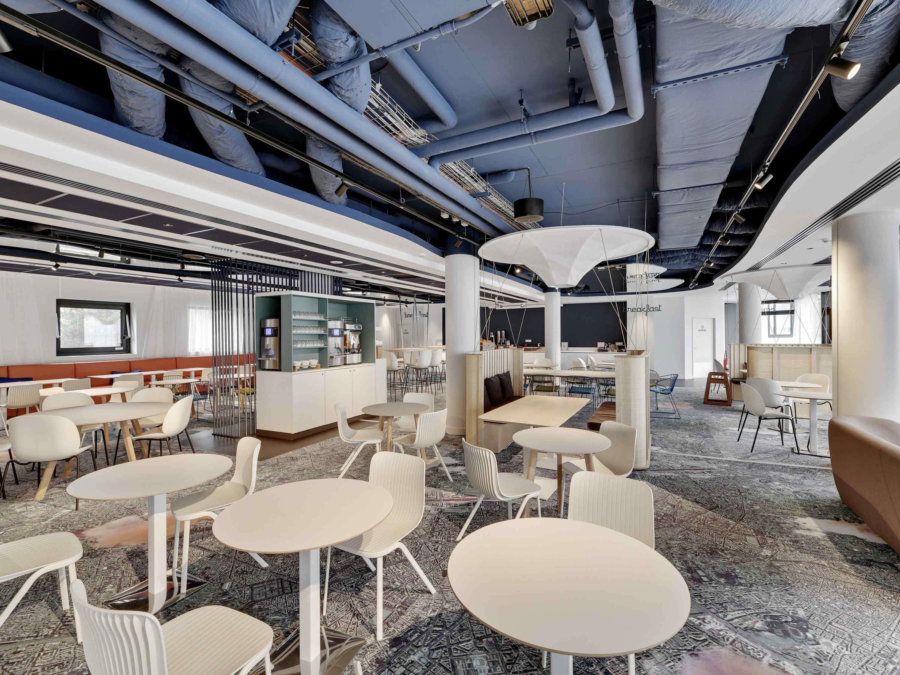 Un área de comedor moderna con mesas y sillas redondas, una estación de café y vistas del techo con tuberías a la vista.