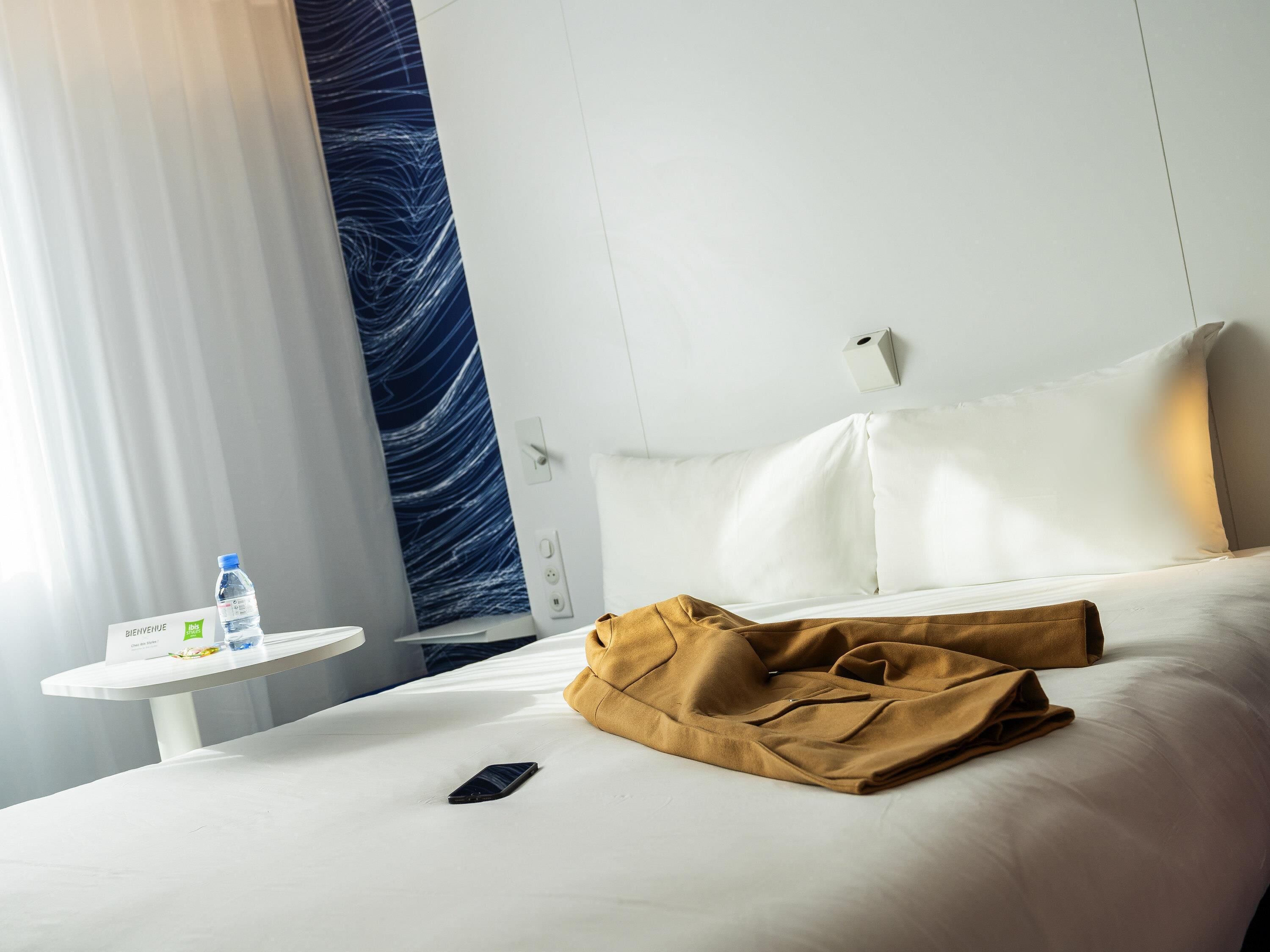 A hotel room with a bed, a bedside table with a water bottle, a smartphone, and a welcome sign.