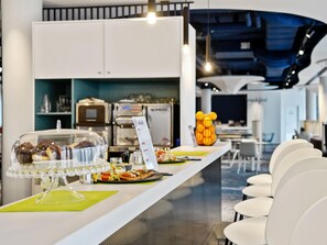 Zona de bar moderna con mostrador que exhibe alimentos, una máquina de café y asientos con sillas blancas.
