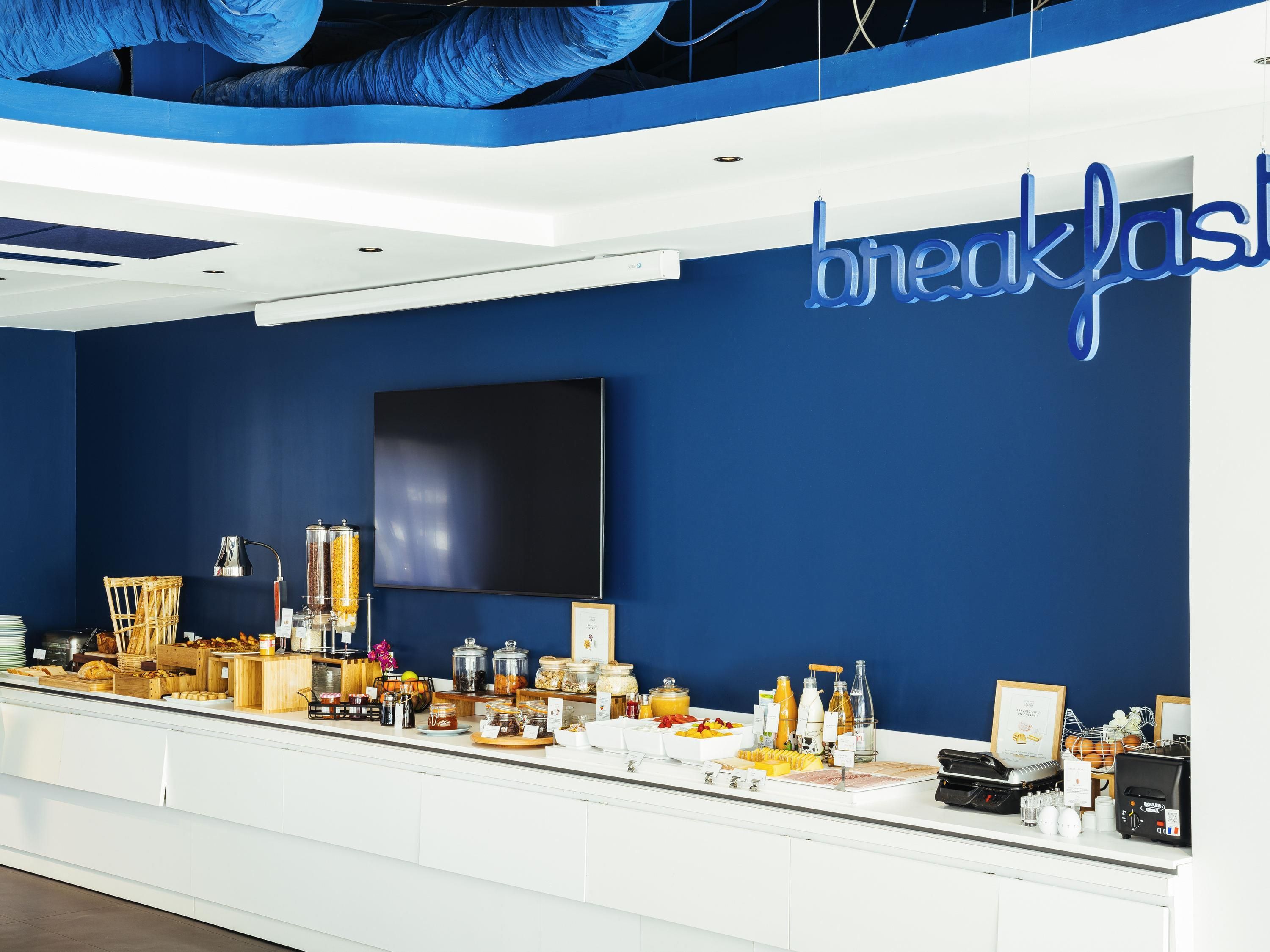 A breakfast buffet with various food items and a large TV screen on the wall.