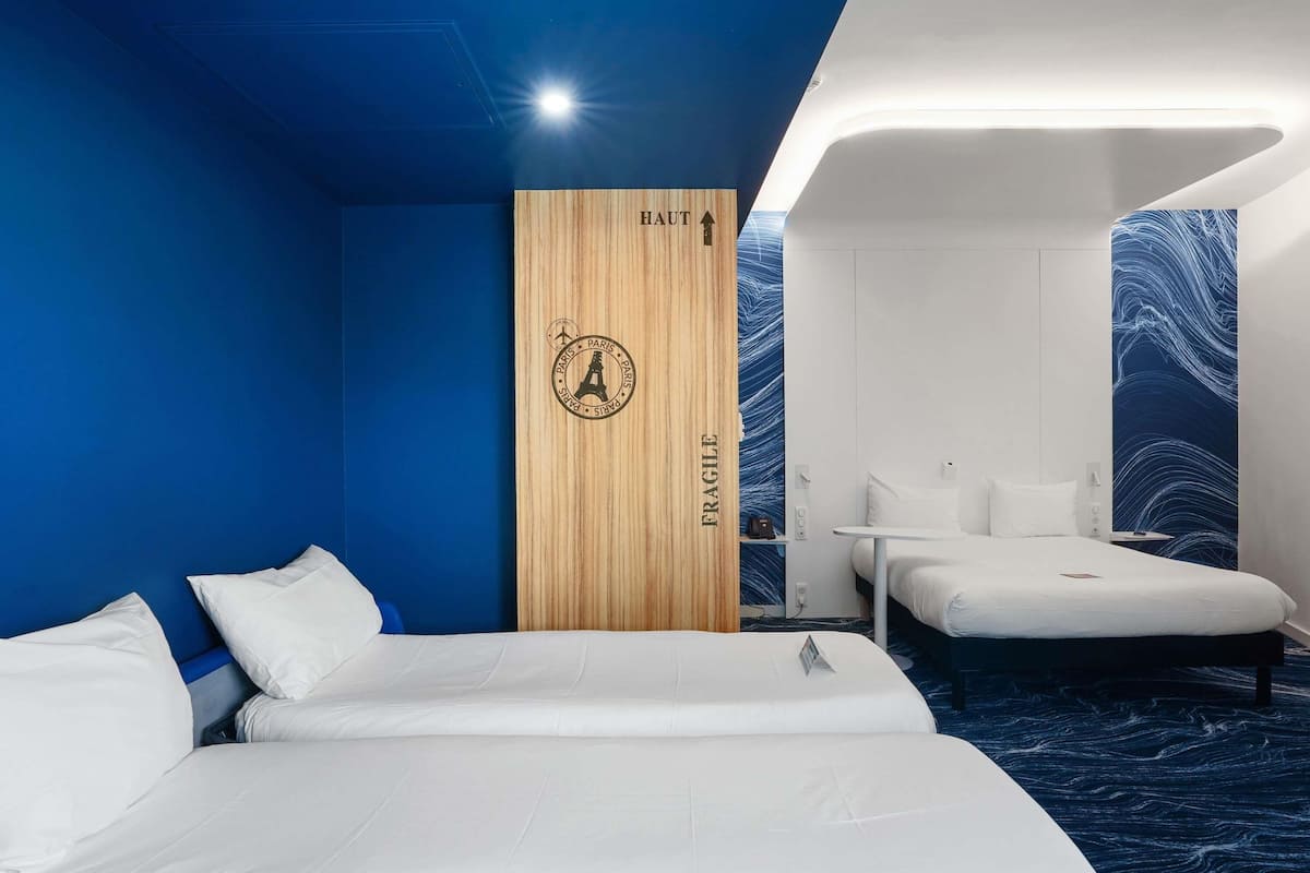 A hotel room with two beds, a wooden partition with 'FRAGILE' and 'PARIS' signage, a small table, and a blue patterned wall.