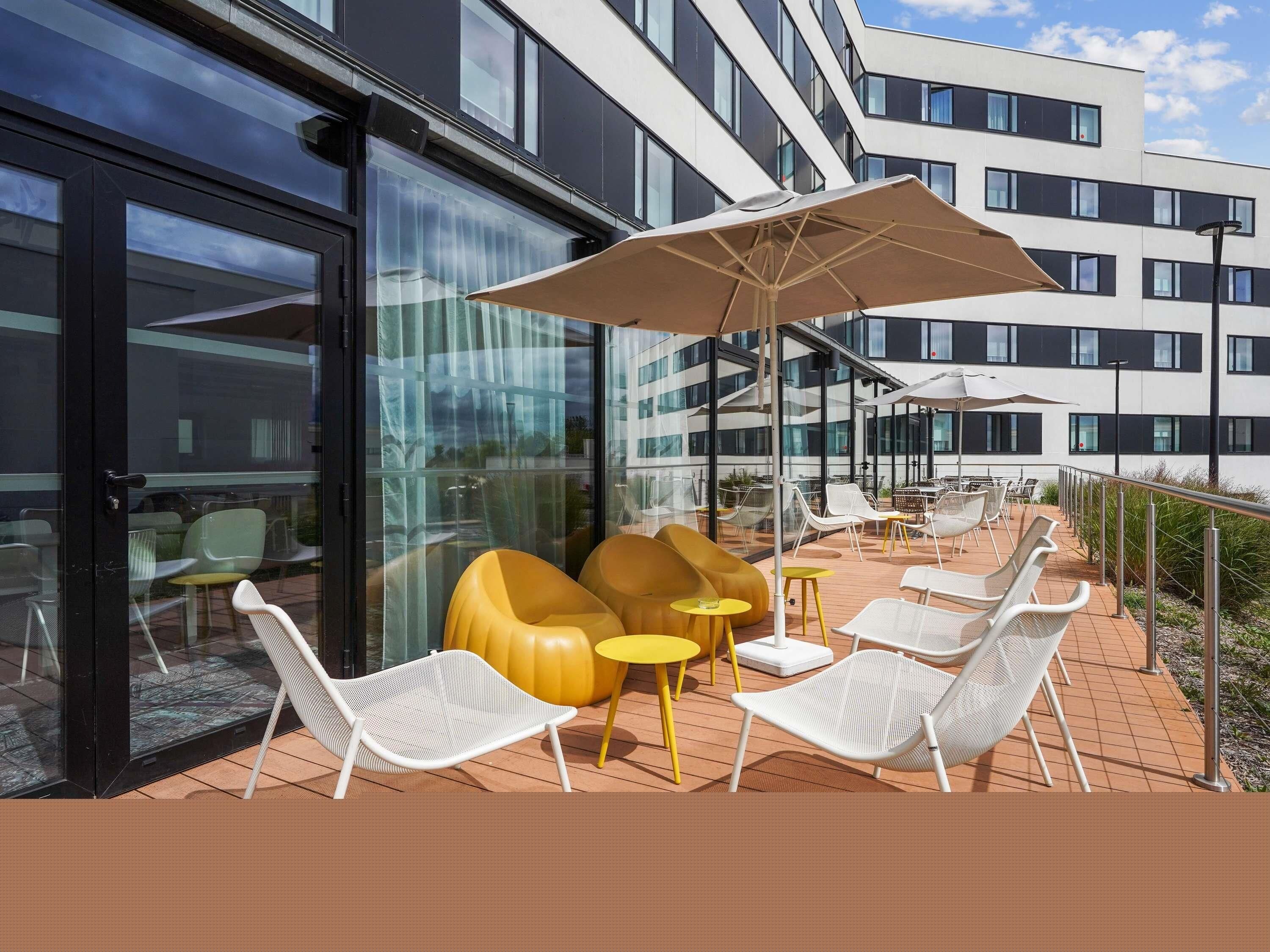 Un espace extérieur aménagé avec des chaises et des tables, un grand parasol, et un bâtiment aux grandes fenêtres en arrière-plan.
