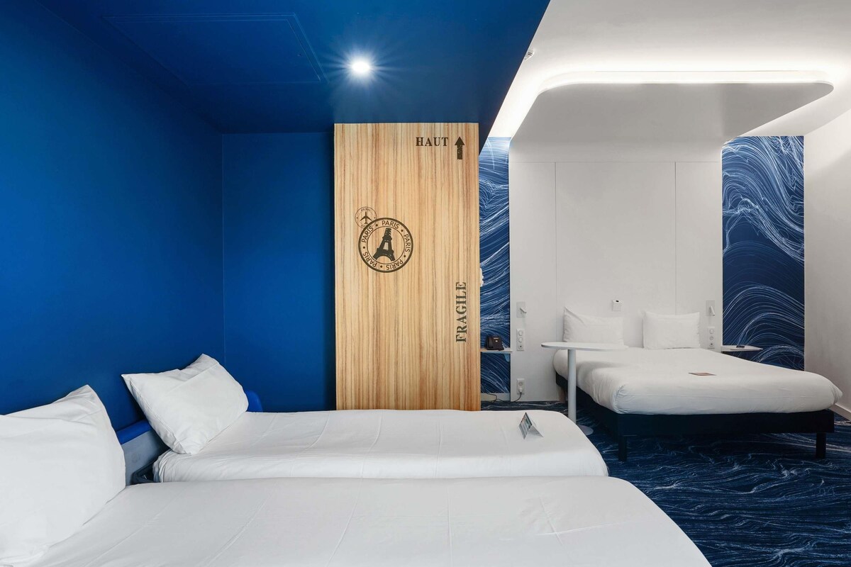 A hotel room with two beds, a wooden partition with 'FRAGILE' and 'PARIS' signage, a small table, and a blue patterned wall.