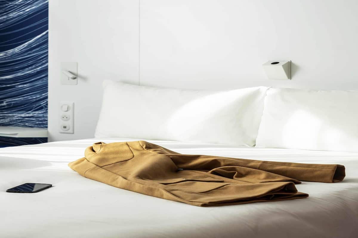 Una cama bien hecha con sábanas blancas, un teléfono inteligente sobre la cama y una prenda marrón en la cama.