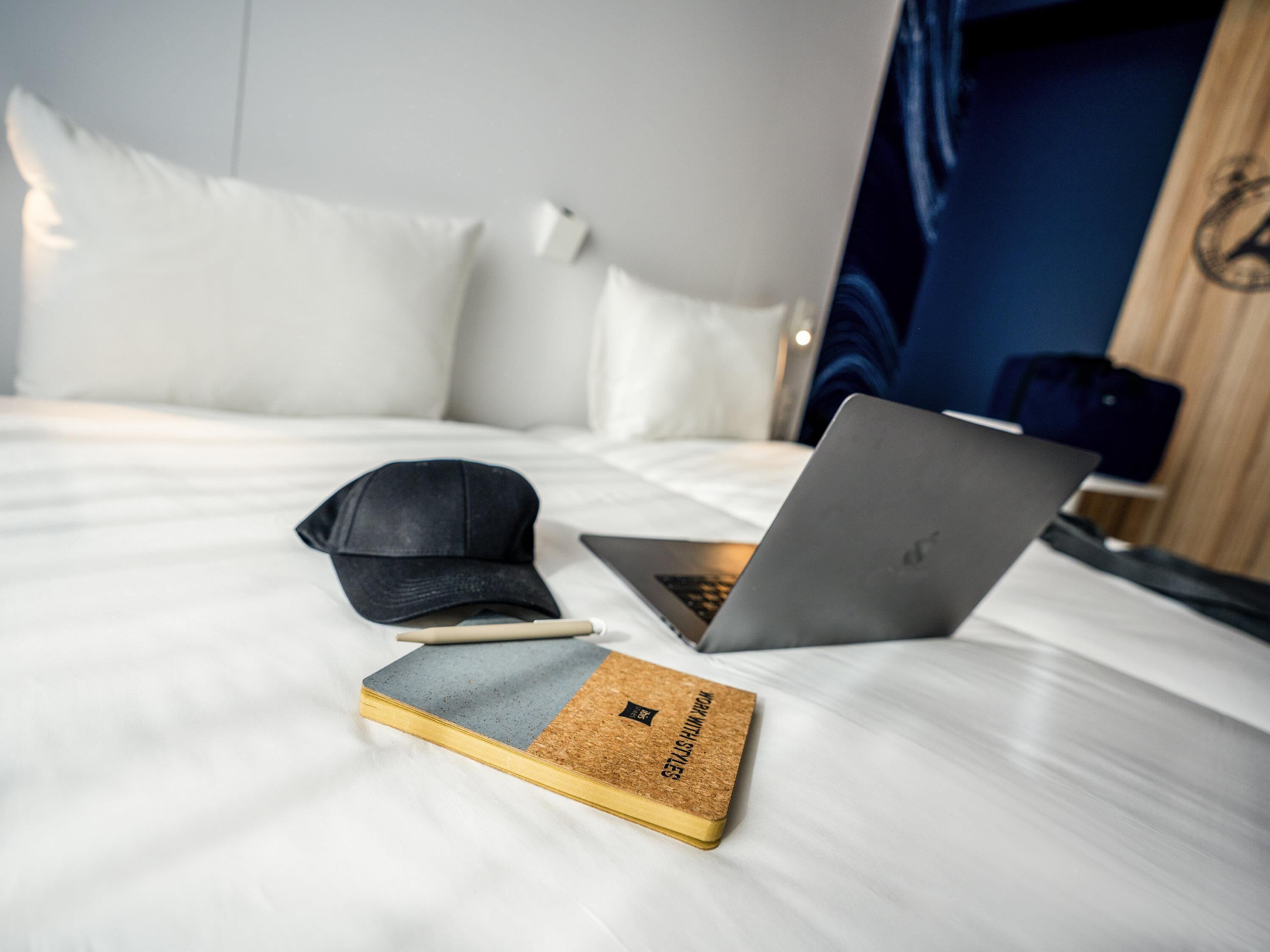 A bed with a laptop, a cap, and a notebook on it.