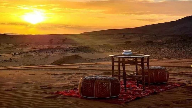Paisaje desértico con una mesa y dos otomanas sobre una tela roja, una mesita con una maceta y un atardecer al fondo.
