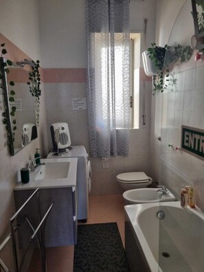 A compact bathroom with a sink, toilet, and bathtub. There is a window with a sheer curtain, and a sign on the wall.