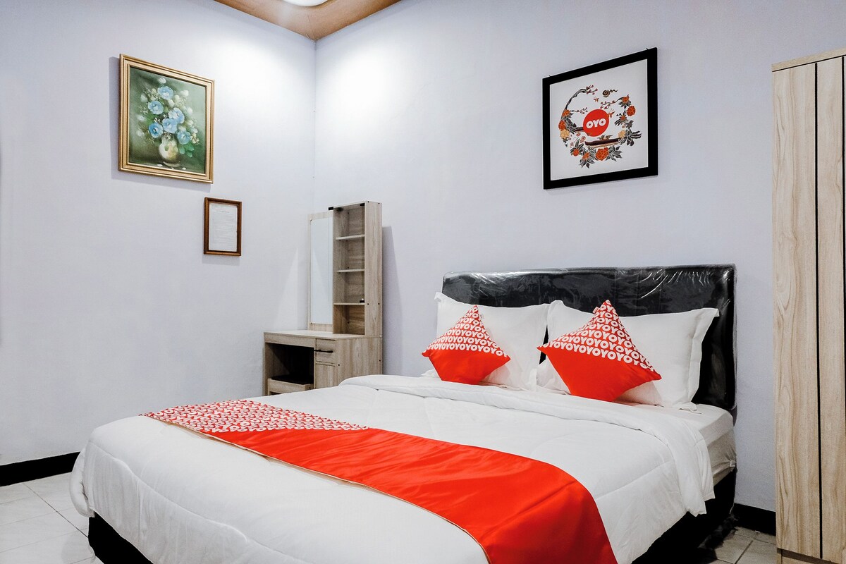 Un lit bien fait, recouvert d’une couvre-lit blanche et rouge, avec deux oreillers rouges et une tête de lit noire. Sur le mur, à gauche, on aperçoit une œuvre d’art florale encadrée, et à droite, une photographie encadrée aux motifs rouges et orange.