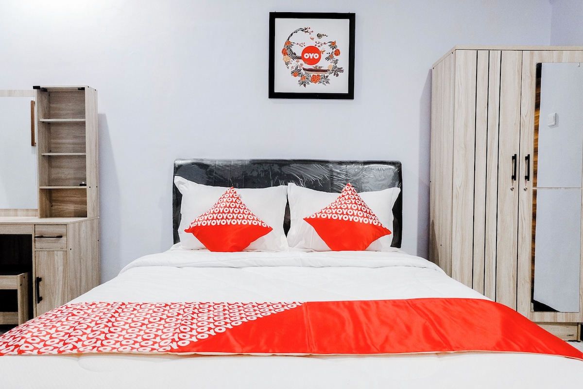 Ein Bett mit weißroter Bettdecke, zwei rote Kissen, ein Holzschrank und ein gerahmtes Bild an der Wand.