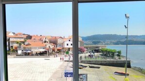 A room with a large window overlooking a beach area with buildings, a promenade, and boats. - Studio with sea views (APT 1) (Combarro)