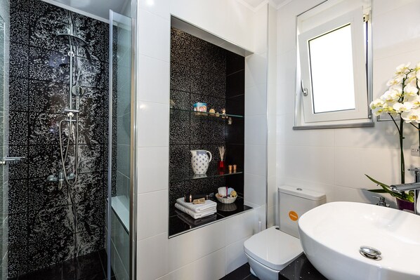 A modern bathroom with a black and white marble shower, a glass shelf with toiletries, a white toilet, and a large white sink.