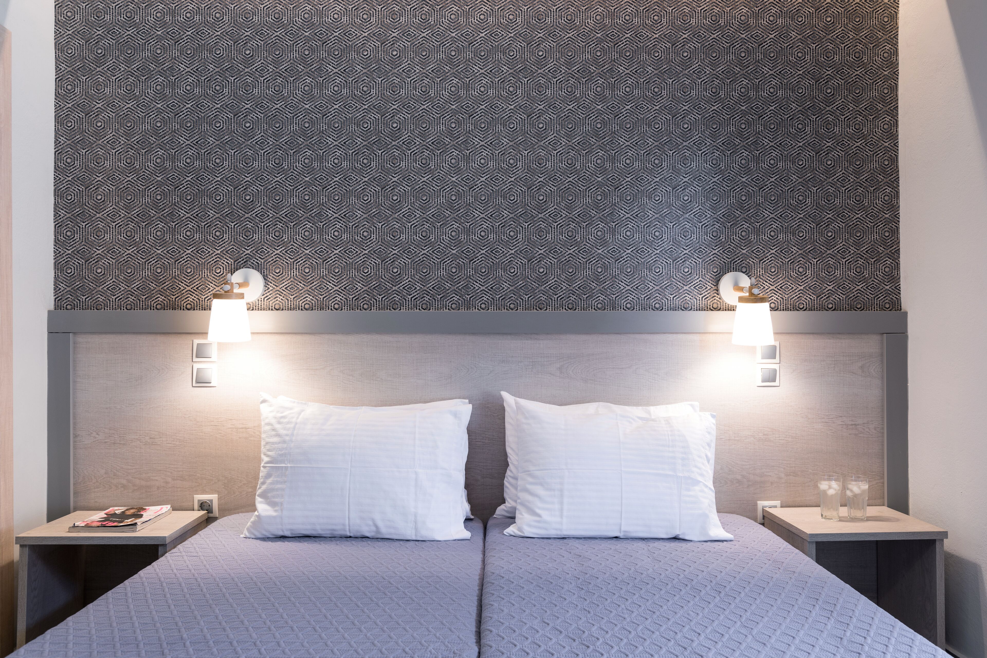 Uma cama de casal com colcha cinza texturizada, duas almofadas brancas e cabeceira com padrão geométrico. Há duas mesas de cabeceira, cada uma com uma luminária e uma revista.