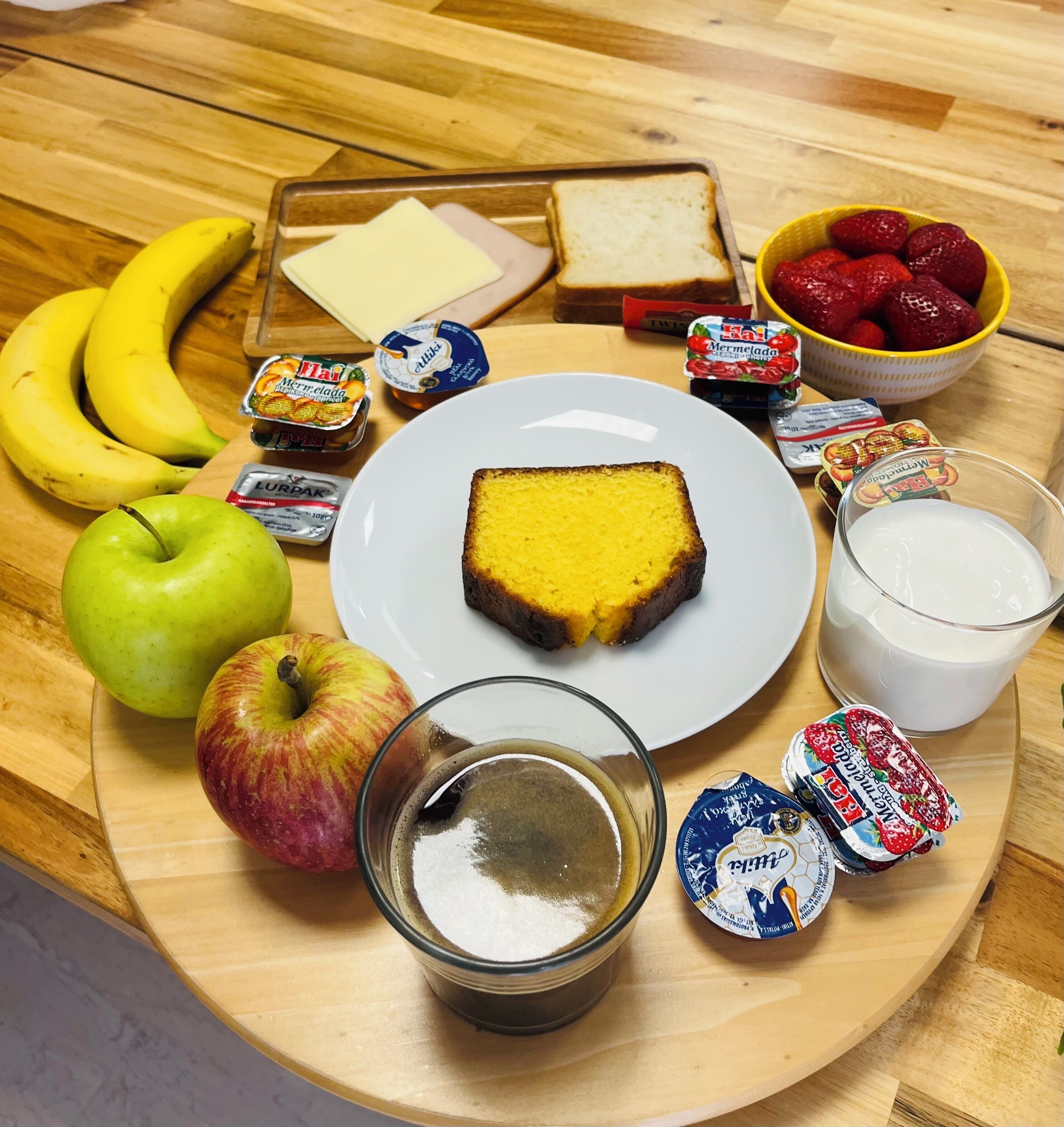 A breakfast tray with a slice of cake, a glass of milk, a bowl of strawberries, a banana, an apple, and various condiments.