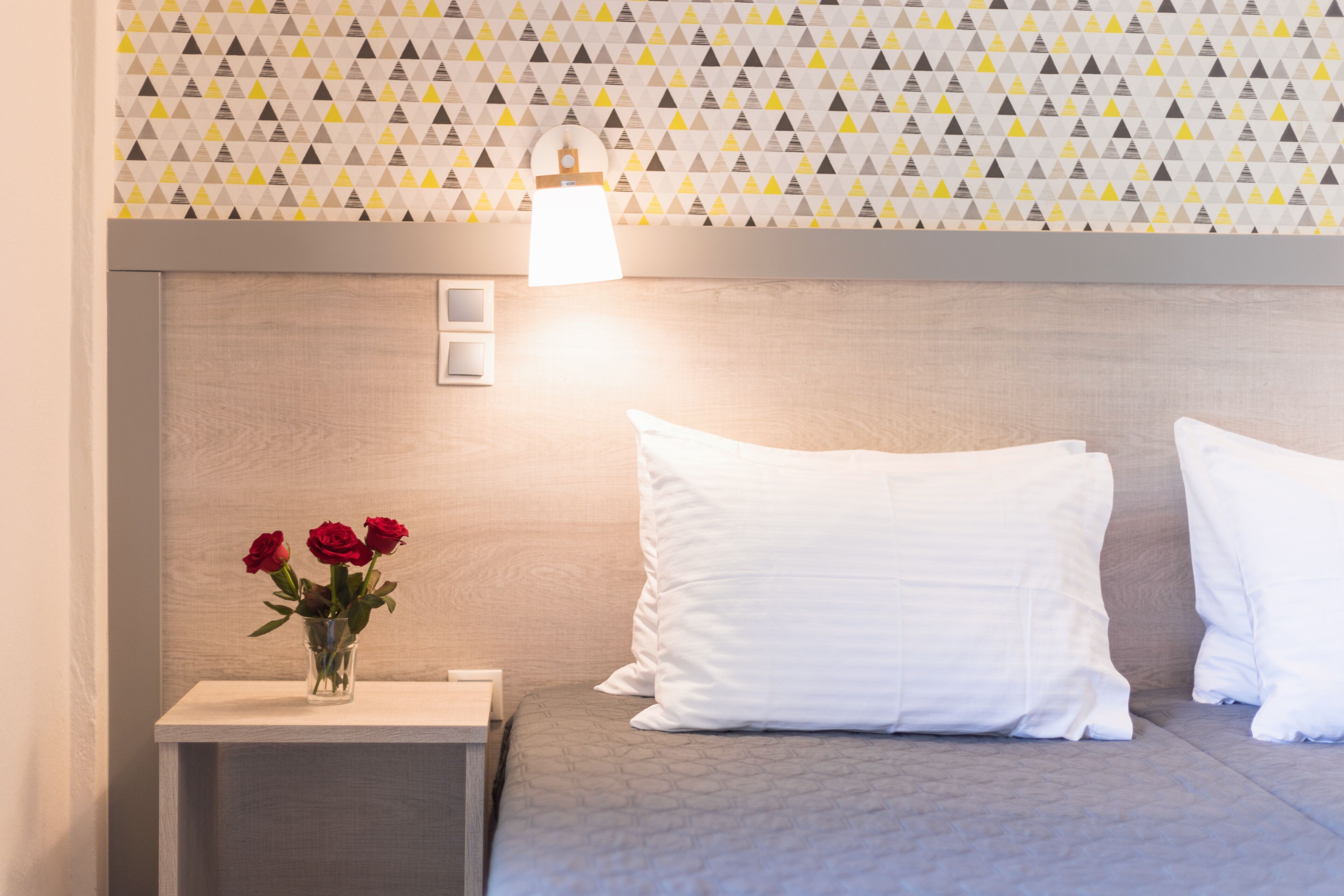 Una cama bien tendida con almohadas blancas, una mesita de noche con un jarrón de rosas rojas y una pared con un diseño geométrico.