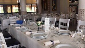 Une salle à manger où les tables sont dressées pour un événement formel, avec des nappes blanches, de la vaisselle, des verres et des couverts disposés avec soin.