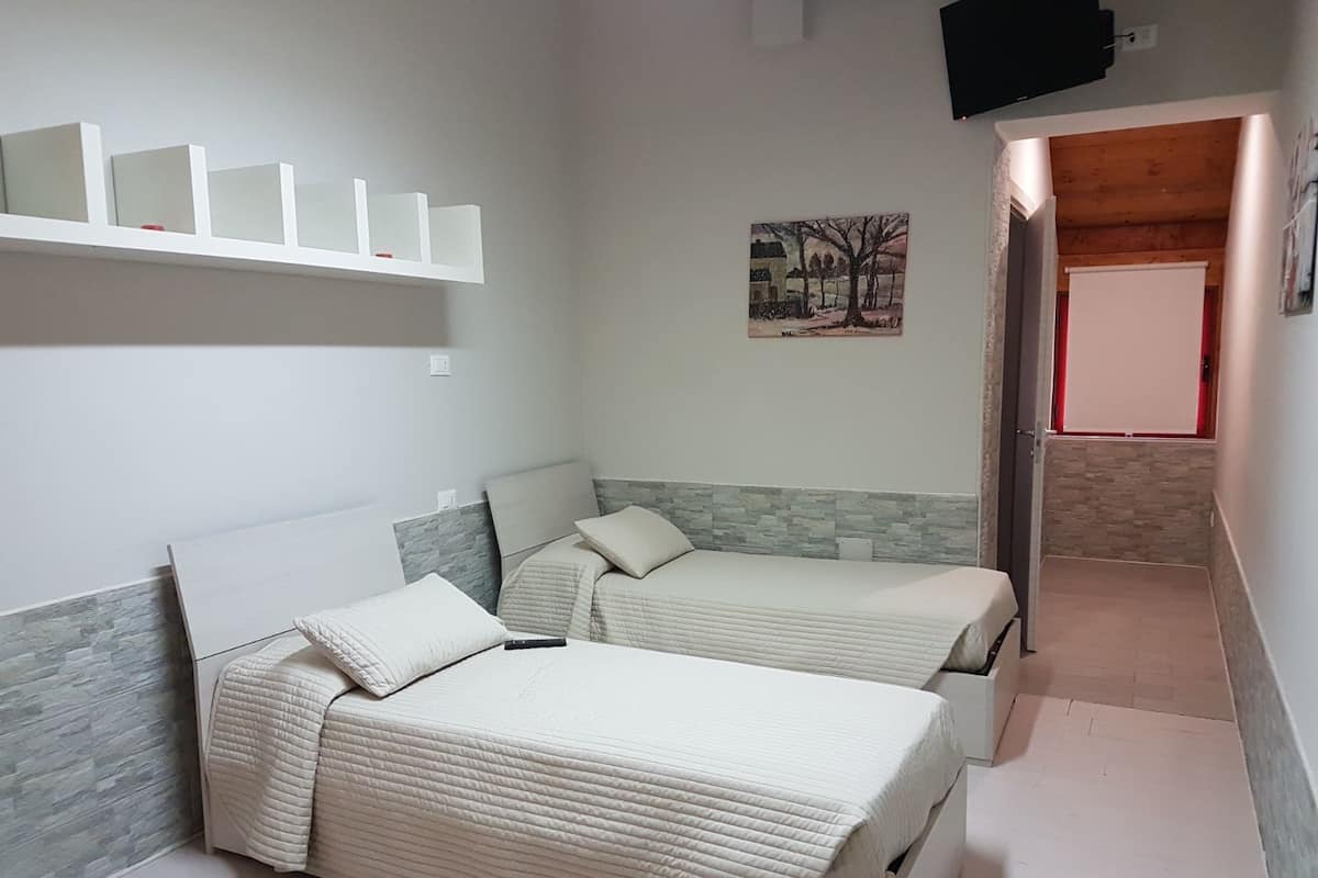 A bedroom with two beds, a mounted TV, a painting, and a small shelf.