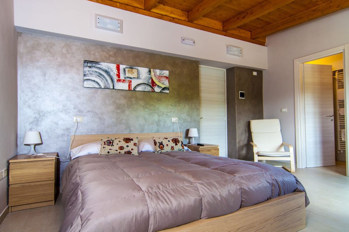 A bedroom with a large bed, wooden headboard, two bedside tables with lamps, a chair, and a door leading to another room.