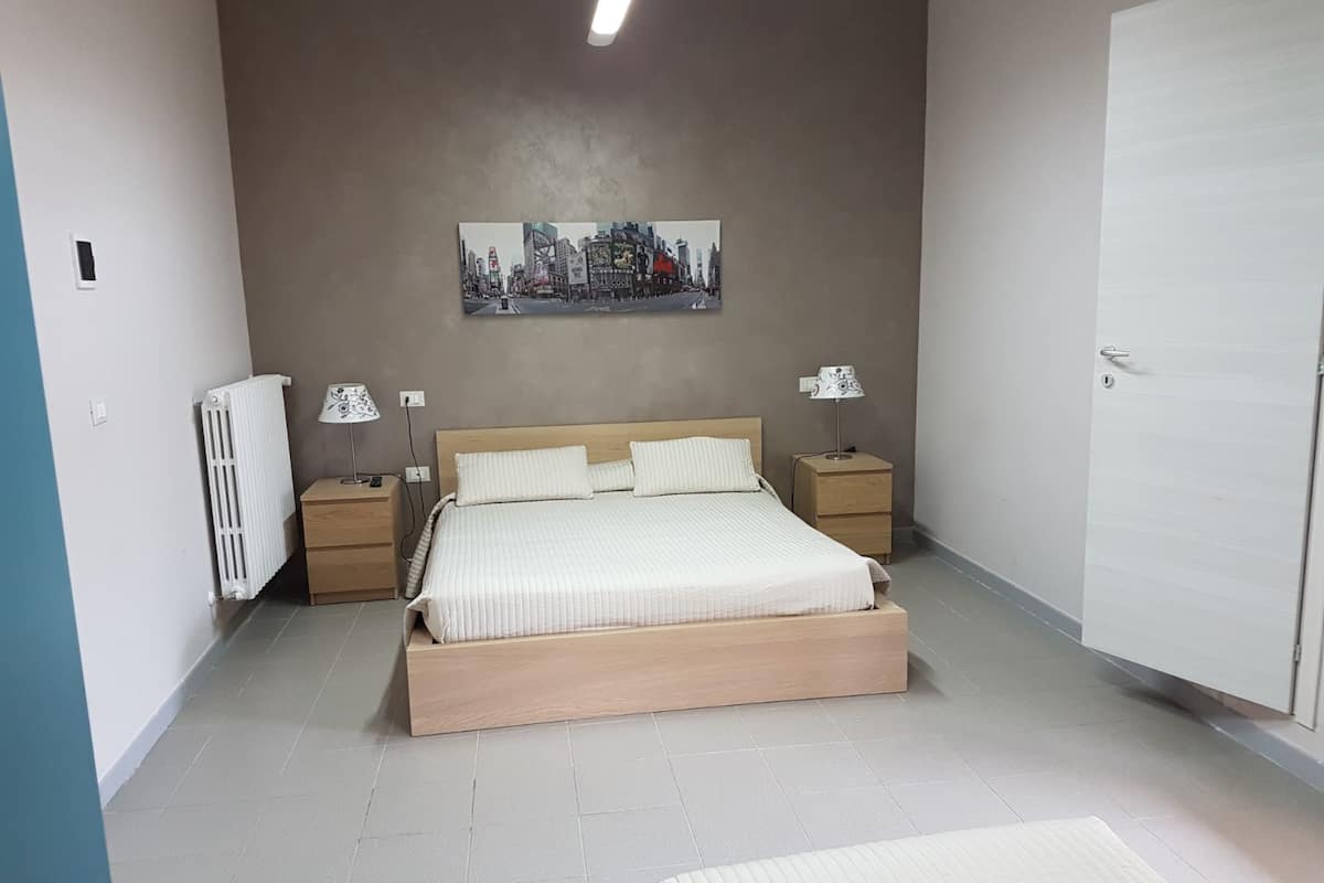 A bedroom with a bed, two bedside tables, a radiator, a wall-mounted painting, and a door.