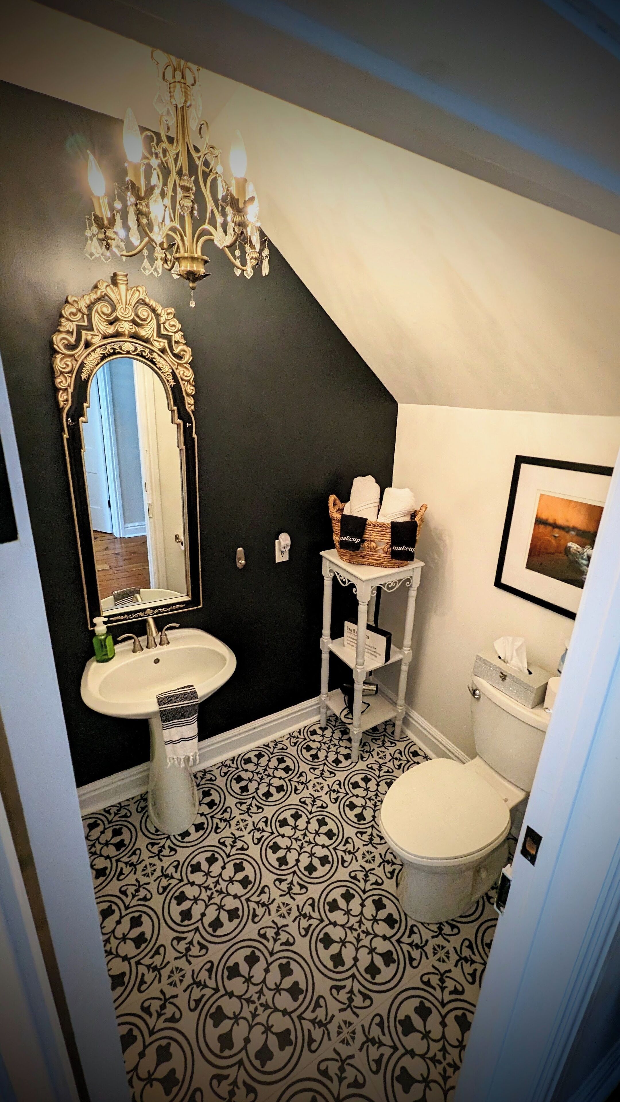 Une salle de bain avec un carrelage noir et blanc, un lavabo blanc avec un miroir doré, des toilettes blanches et une petite commode blanche surmontée d’un panier rempli de serviettes.