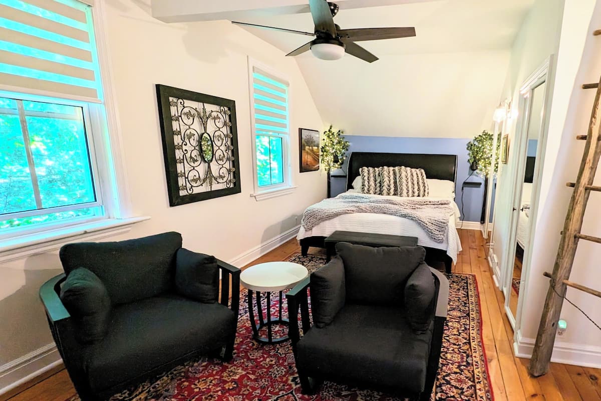 A bedroom with a bed, two armchairs, a small round table, a ceiling fan, and a wooden ladder.