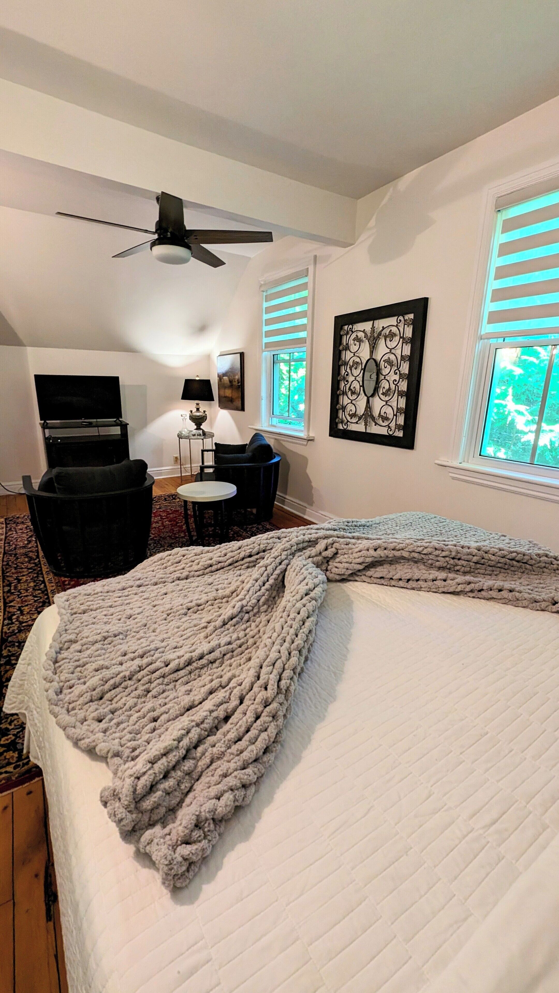 A hotel room with a bed, a desk, a chair, a ceiling fan, and a window with blinds.