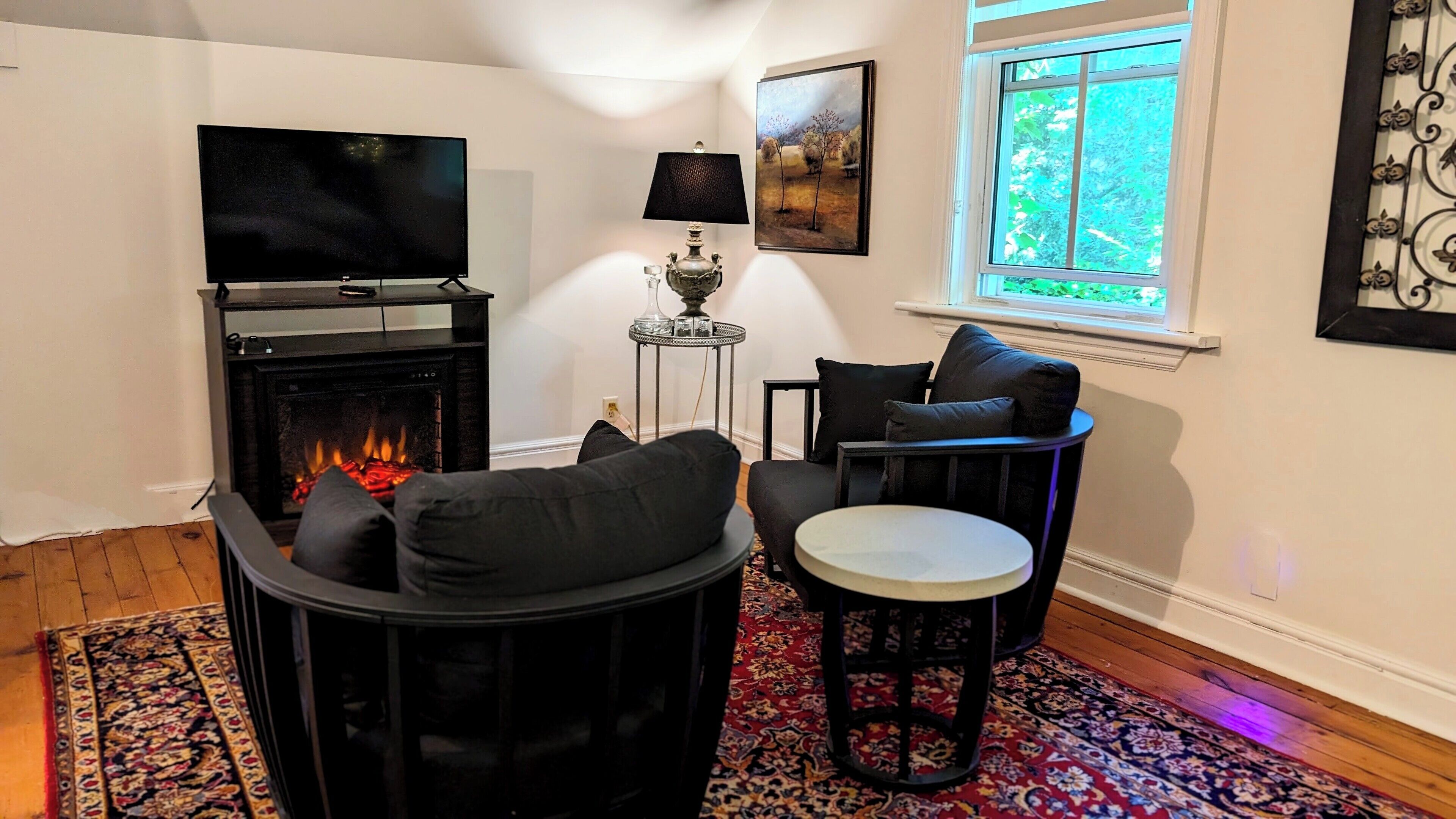 A living room with a fireplace, a flat-screen TV, a table, and a window with a view of greenery.
