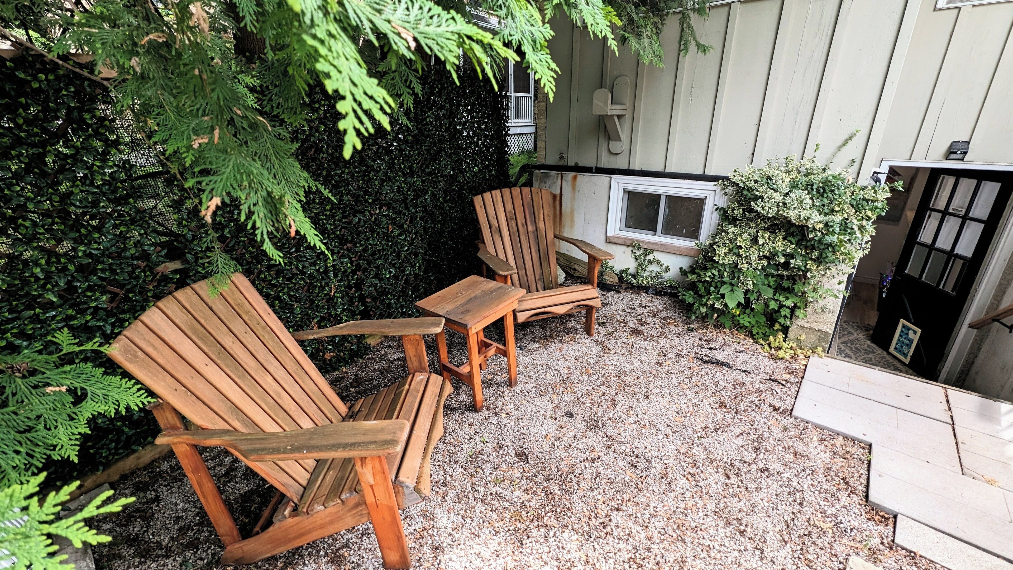 A patio with two wooden Adirondack chairs and a small table, surrounded by gravel and greenery.