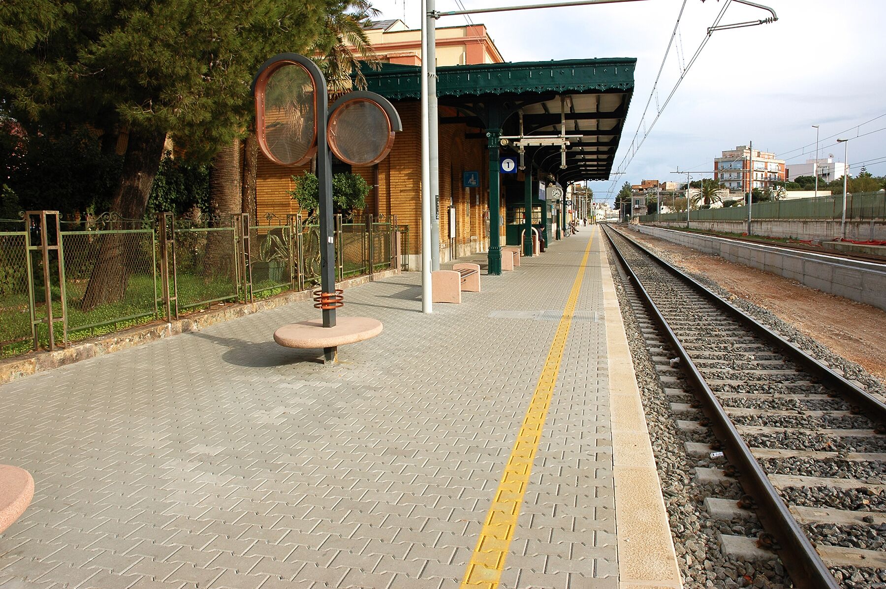 Ein Bahnsteig mit überdachtem Wartebereich, Bänken und einem Zaun.