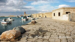 Eine Uferpromenade mit vor Anker liegenden Booten, ein Leuchtturm in der Ferne und eine Steinmauer mit Rundbögen.