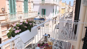 Ein Balkon mit einem weißen Metalltisch, gedeckt mit einem Teekessel, Tassen und Gebäck. Im linken Bereich befindet sich ein Pflanzgefäß mit rosa Blumen.