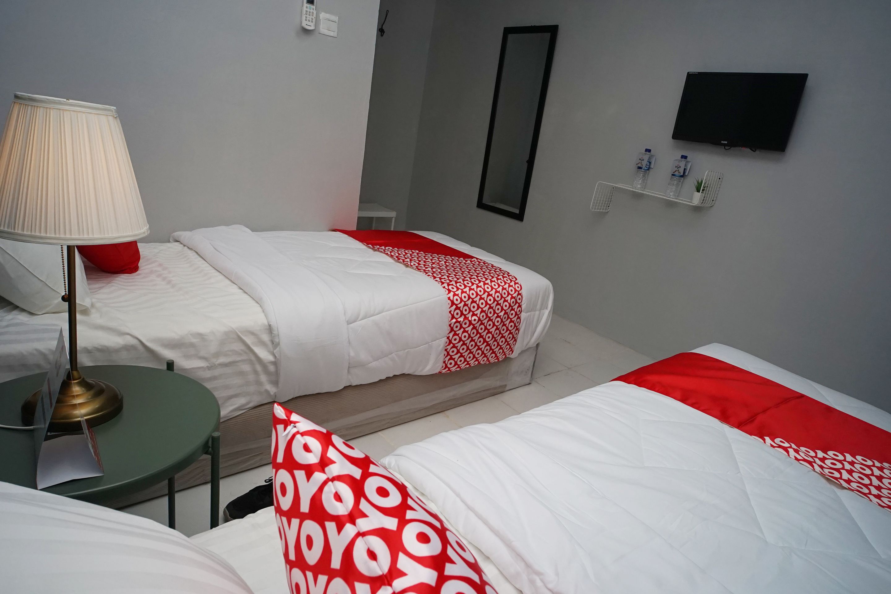 A hotel room with two beds, a bedside table with a lamp, a mirror, a TV, and a shelf with water bottles.