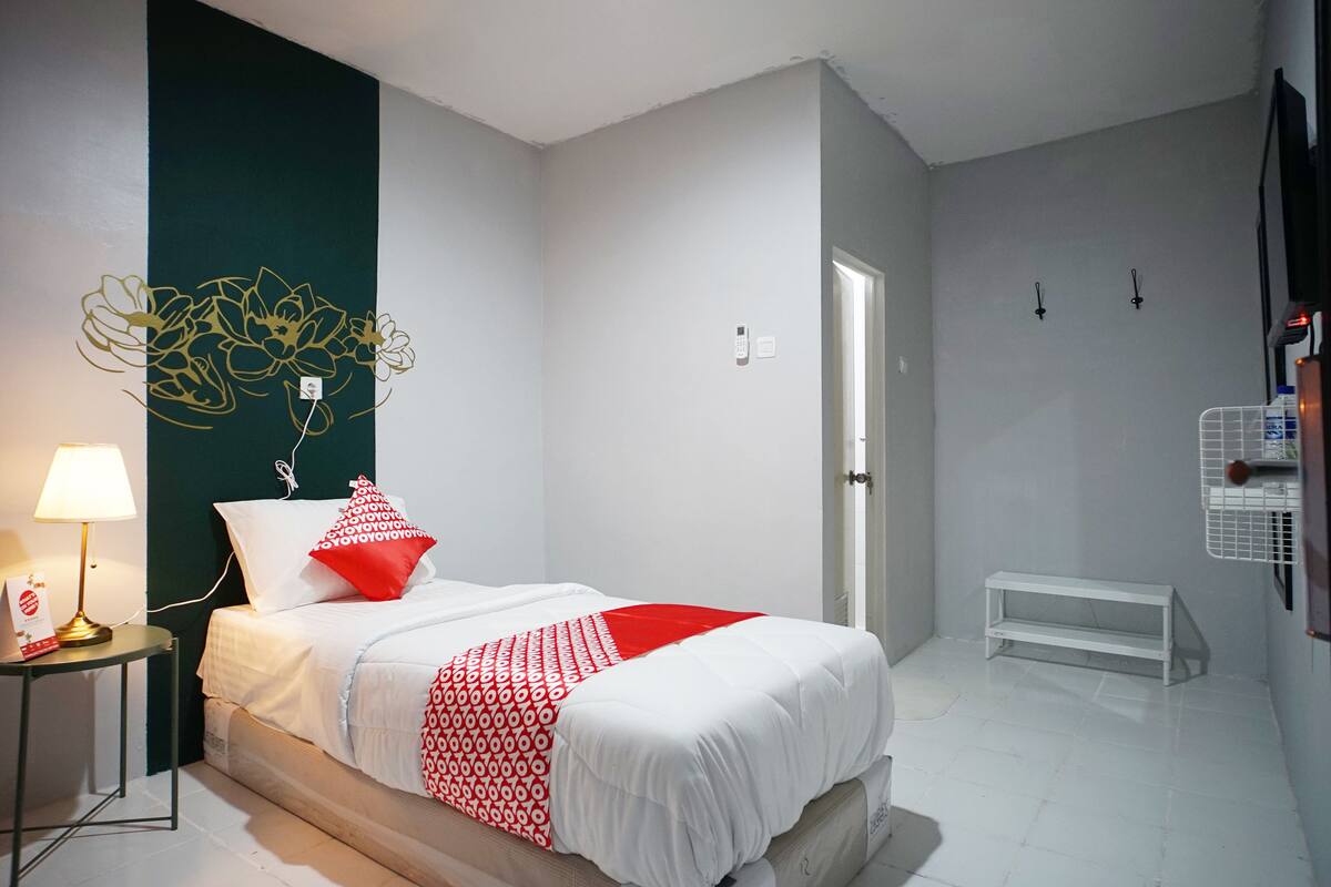 A bedroom with a bed, a bedside table with a lamp, a wall art piece, a door, and a white bench.