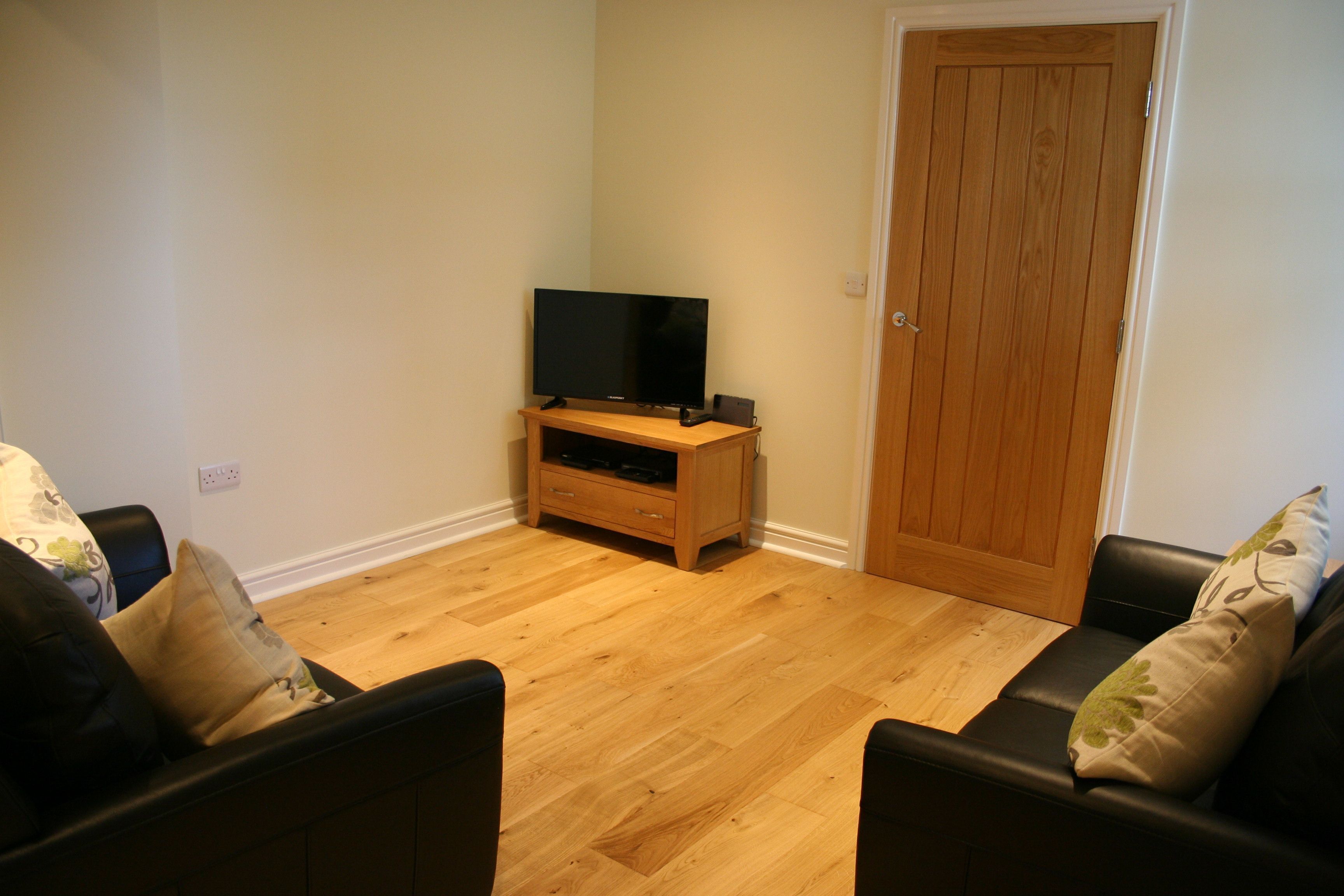 A living room with a wooden floor, a television on a wooden stand, and a door in the background.