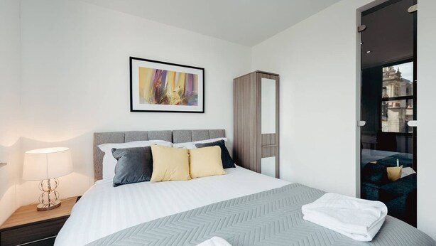 A modern hotel room with a large bed, a bedside table with a lamp, a wardrobe, and a framed abstract painting on the wall.