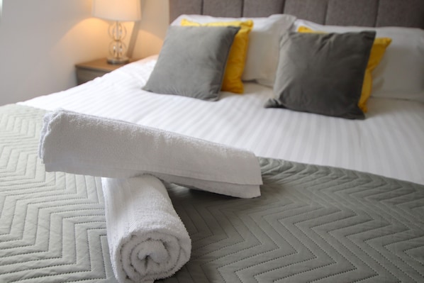 A neatly made bed with a rolled white towel, grey and yellow pillows, and a grey quilted bedspread. - The Ridings Apartment 214 (Halifax)