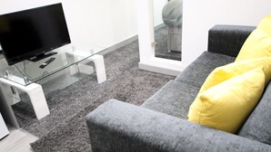 A modern living room with a flat-screen TV on a glass coffee table, grey sofa with yellow cushions, and a grey carpet.