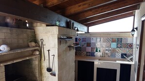Uma cozinha rústica com um forno de tijolos, prateleiras com utensílios de cozinha, um revestimento de azulejos e uma janela.