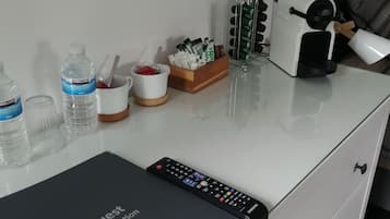 A hotel room with a white desk, a black remote control, a coffee maker, a water bottle, a mug, and a small box of tissues.