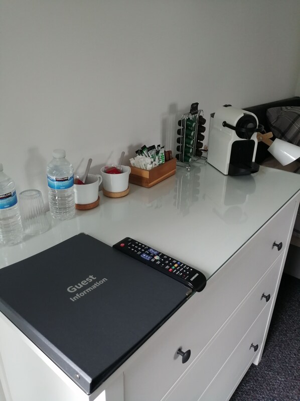 Una habitación de hotel con un escritorio blanco, un control remoto negro, una cafetera, una botella de agua, una taza y una pequeña caja de pañuelos.