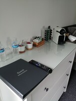 Una habitación de hotel con un escritorio blanco, un control remoto negro, una cafetera, una botella de agua, una taza y una pequeña caja de pañuelos.