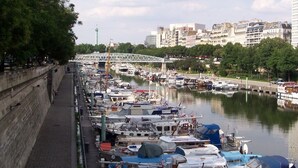 Un port de plaisance où de nombreux bateaux sont amarrés le long d’une promenade en bord de mer, un pont en arrière-plan et des bâtiments de l’autre côté de la rivière.
