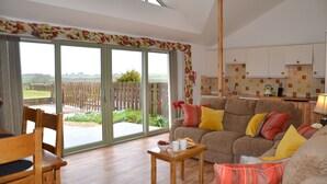 A spacious living room with a large window, a wooden dining table, and a sofa with colorful cushions. - Willow Cottage Purpose built wheelchair friendly cottage (Truro)