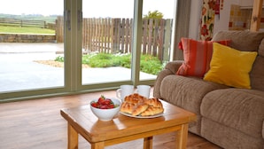 A wooden table with a bowl of strawberries and croissants, a sofa with colorful pillows, and a view of a garden through a glass door. - Willow Cottage Purpose built wheelchair friendly cottage (Truro)