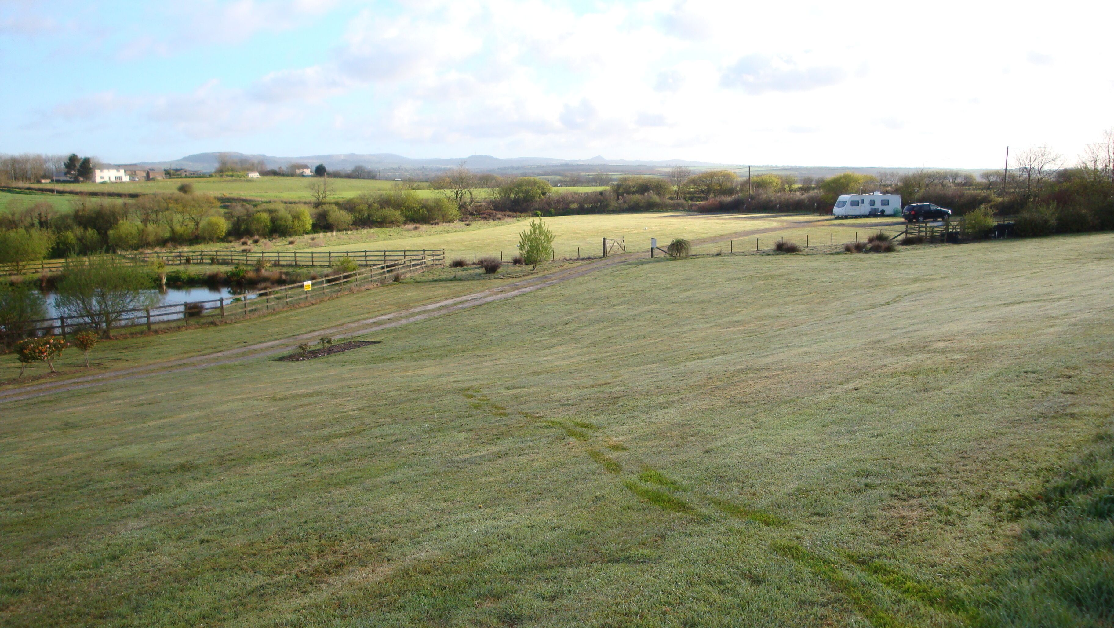 A rural landscape with a field, a small body of water, a white caravan, and distant hills.