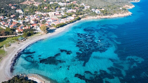 An aerial view of a coastal town with buildings along the shoreline, clear blue waters, and a hilly backdrop.
