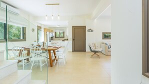 Un comedor moderno y bien iluminado, con una mesa de madera y sillas blancas, que da acceso a una cocina y a una sala de estar con sofá y sillón.