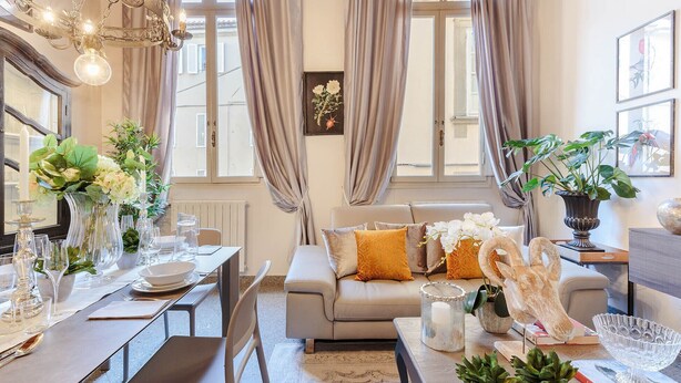A well-lit living room with a dining area, featuring a sofa, dining table set for a meal, and various decorative items. - ARIAS APARTMENT in the iconic Piazza San Michele (Lucca)