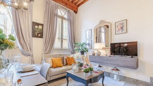 A cozy living room with a sofa, coffee table, and a TV. There are curtains, a chandelier, and a painting on the wall. - ARIAS APARTMENT in the iconic Piazza San Michele (Lucca)