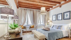 A bedroom with a wooden ceiling, a bed with a blue bedspread, a bench, a desk with a lamp, and a window with curtains. - ARIAS APARTMENT in the iconic Piazza San Michele (Lucca)